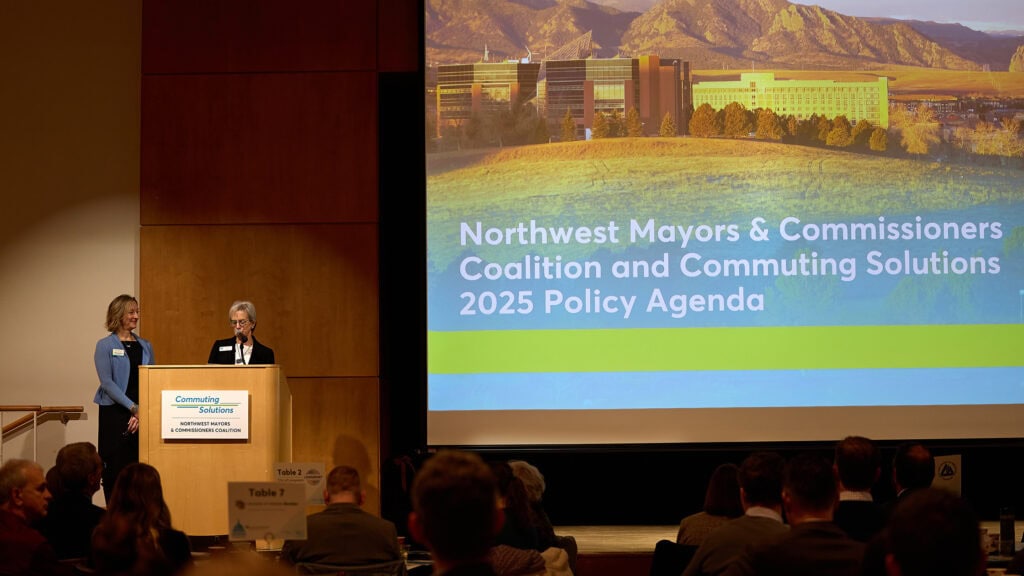 Boulder County Commissioner Claire Levy and Commuting Solutions Board Chair Kirsten Schuchman stand on stage near presentation screen