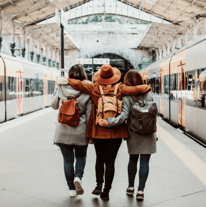 Three women walk with their arms around each other on a train platform