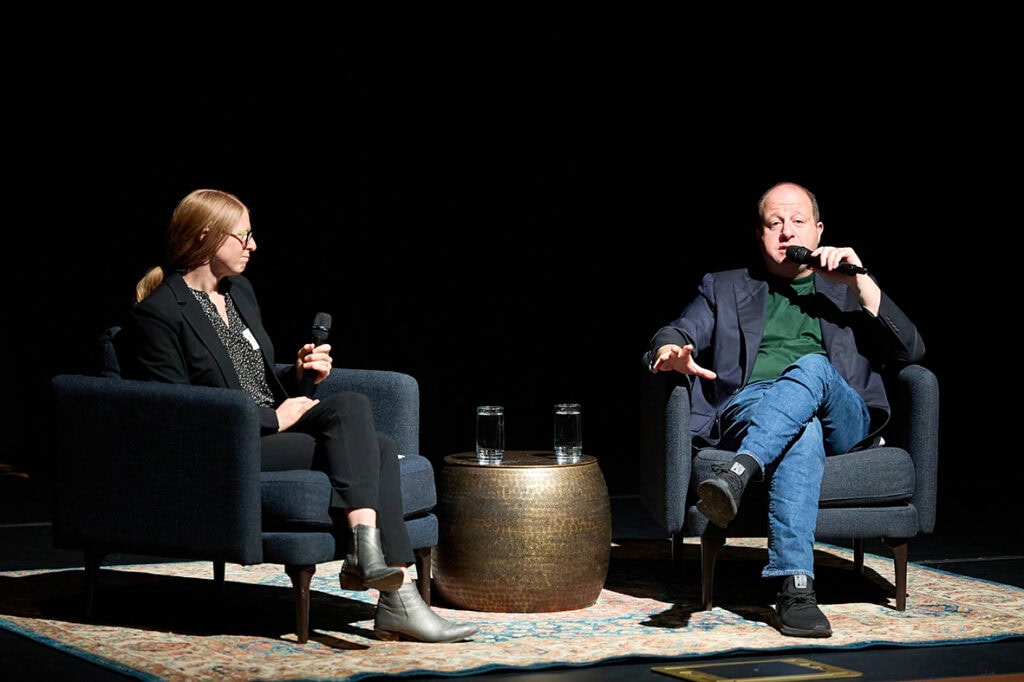 Governor Jared Polis sitting on stage holding mic with female interviewer