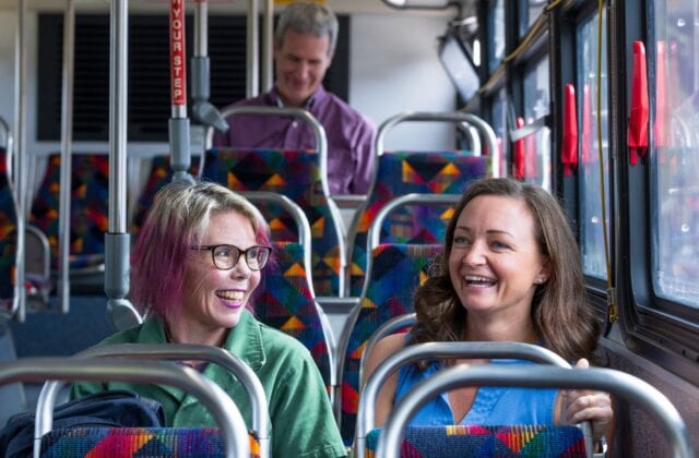 Two people sit in seats on a city bus with smiling faces. Another passenger sits two rows behind them.