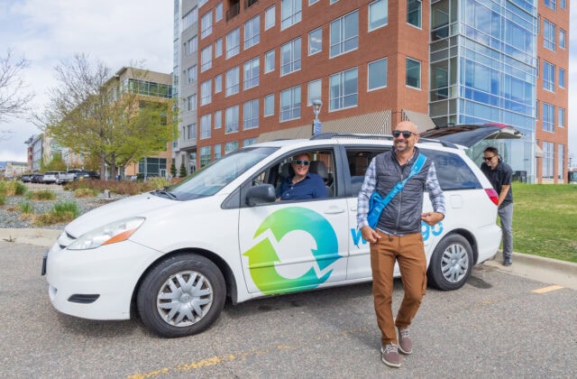 Driver seated in a white Way to Go minivan, which features a green and blue recycle symbol and the words 'Way to Go' on the side. The driver is looking out the window at a commuter who just exited the vehicle, while another person is placing groceries into the van's trunk.