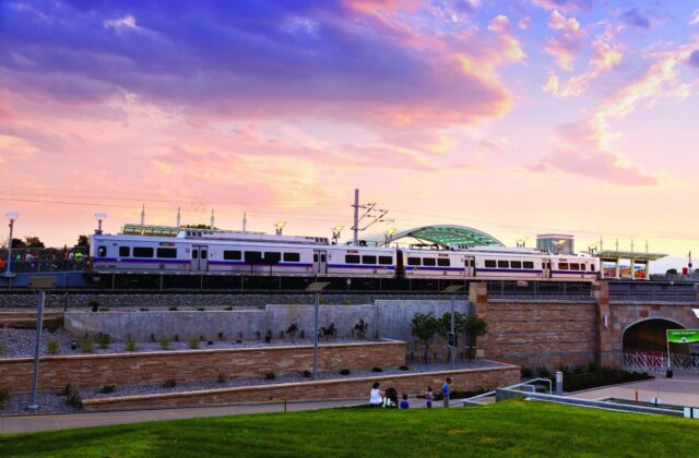 Commuter train sits on a bridge in front of a setting sun over the Rocky Mountains.