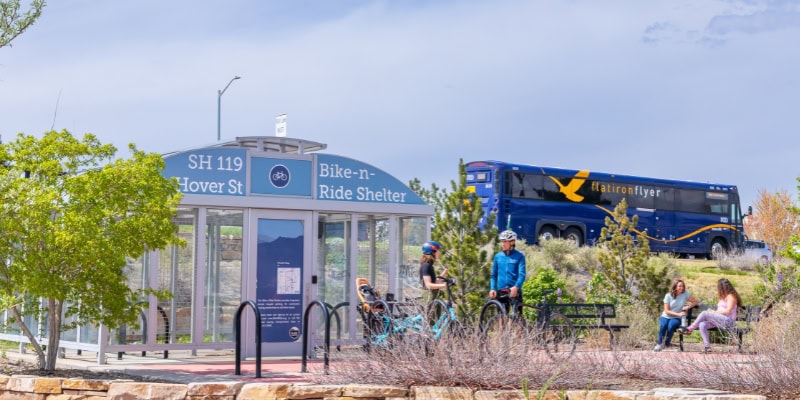 a corner transit stop in Longmont Colorado shows 2 cyclists standing in front of a bike shelter with a bus in the background