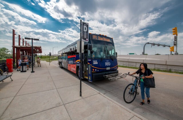 A smiling cyclist walks with her bike to the sidewalk near an RTD bus stop after unloading her bike from the bus.